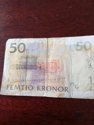 Svensk 50-lapp sedel - Säljer en svensk 50-kronorssedel med motiv av noter, instrument och porträtt. Sedeln har detaljer i gult, lila och grått samt texten FEMTIO KRONOR. Perfekt för samlare eller som present.