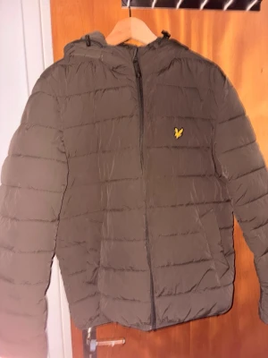 Olivgrön pufferjacka från Lyle & Scott - Säljer en olivgrön pufferjacka från Lyle & Scott med huva och dragkedja framtill. Jackan har det klassiska gula logomärket på bröstet och quiltad design som ger en schysst streetlook. Perfekt för vårens dagar när du vill hålla värmen och ändå se snygg ut