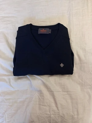 Mörkblå tröja med V-ringning Morris - Klassisk mörkblå tröja från Morris med V-ringning och diskret logga broderad på bröstet. Tröjan har långa ärmar, ribbade muddar och är tillverkad i ett mjukt material som passar perfekt till lager på lager-stil.