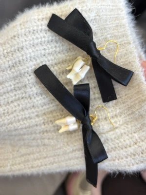 Svarta rosettörhängen med tänder - Unika örhängen med svarta satinrosetter och vita tandformade hängen. Krokarna är guldfärgade och ger en cool kontrast till de svarta rosetterna. Perfekt för dig som vill sticka ut med accessoarer som verkligen syns.