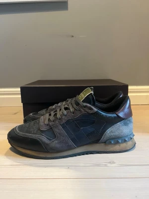 Valentino Garavani sneakers grå/svart - Snygga sneakers från Valentino Garavani i grått och svart med detaljer i mocka, läder och textil. Skorna har nitar på hälen och en beige sula som ger en cool kontrast. Perfekta för dig som gillar exklusiva och unika sneakers.