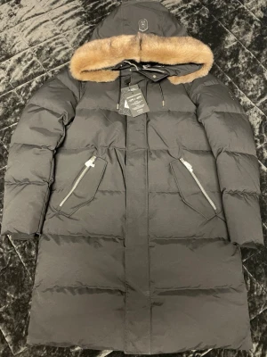 Svart lång dunjacka med pälskant från MACKAGE - Snygg svart lång dunjacka från Moorer med stor huva och lyxig pälskant i beige. Jackan har två snedställda fickor med dragkedja och quiltad design. Perfekt för kalla vinterdagar och riktigt varm tack vare dunfyllningen.