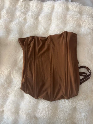 Brun korsett med snörning - Säljer en snygg brun korsett med vertikala sömmar och snörning i ryggen. Korsetten har en mjuk, glansig finish och breda band för justering. Perfekt för att skapa en markerad midja och trendig siluett. De står xl i toppen men skulle säga att den är mindre än så