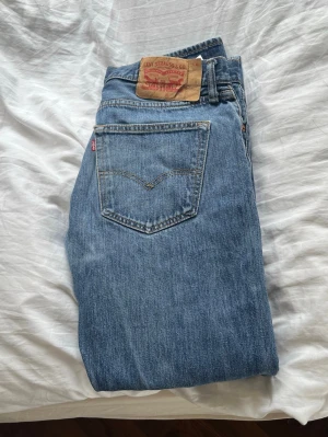 Levi's 501 - Klassiska blå jeans från Levi's med rak passform och femficksmodell. Jeansen har den ikoniska läderlappen bak i midjan och röd Levi's-tagg på bakfickan. Tillverkade i slitstark denim, perfekta för en avslappnad och tidlös stil. Modell: 501