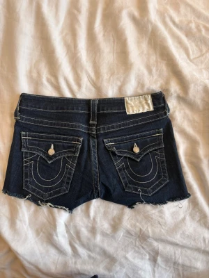 Shorts - Säljer ett par mörkblå jeansshorts från True Religion med råa, fransiga kanter och kontrastsömmar. Shortsen har fickor både fram och bak med dekorativa knappar.
