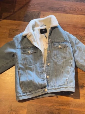 Ljusblå jeansjacka med foder från BikBok - kan alltid diskutera pris! Snygg ljusblå jeansjacka från BikBok med vitt teddyfoder på insidan och kragen. Klassisk modell med två bröstfickor med lock och silverfärgade knappar framtill. Perfekt för dig som vill ha en cool och bekväm jacka med extra värme.