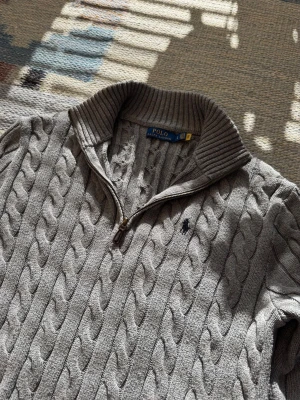 Grå kabelstickad tröja Polo Ralph Lauren - Snygg grå kabelstickad tröja från Polo Ralph Lauren med halv dragkedja och hög krage. Tröjan har ribbade muddar och nederkant samt den klassiska broderade loggan på bröstet. Perfekt för lager på lager och en stilren look.