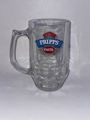 Pripps Fatöl ölsejdel i glas - Klassisk ölsejdel i tjockt glas med Pripps Fatöl-logga i blått och rött. Sejdeln har ett rejält handtag och mönstrad yta som ger ett coolt grepp. Perfekt för dig som gillar att samla på ölglas eller vill ha en unik sejdel till hemmabaren.
