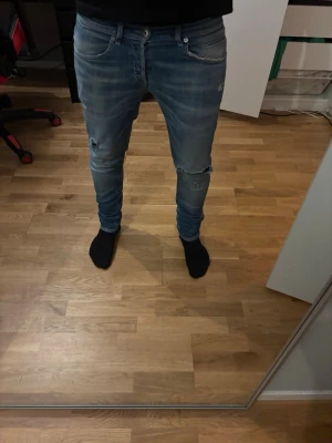 Dondup George - Säljer ett par slim fit ljusblå jeans med snygga slitningar på knäna och klassisk femficksdesign. Jeansen har en smal passform och är tillverkade i denim med lite stretch för extra komfort. Perfekta för en trendig och avslappnad stil.