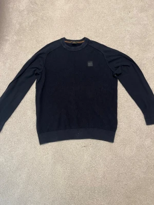 Mörkblå stickad tröja från BOSS - Snygg mörkblå stickad tröja från BOSS med diskret logopatch på bröstet. Tröjan har rund halsringning och ribbade muddar vid ärmslut och nederkant. Perfekt för dig som gillar stilrena och klassiska plagg.