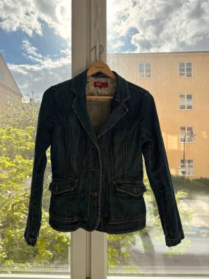 Mörkblå jeansjacka med fickor - Snygg mörkblå jeansjacka från Odd Molly med figurnära passform, två framfickor med lock och dekorativa sömmar. Klassisk krage och knäppning framtill. Perfekt för dig som vill ha en unik twist på den klassiska jeansjackan.