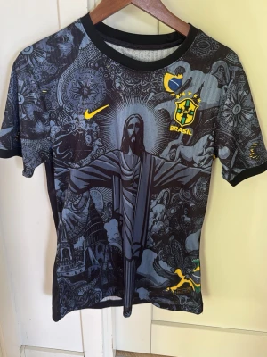 Brazil Ronaldinho Nike fotbollströja - Snygg svart Brasilien fotbollströja från Nike med unikt grafiskt tryck av Kristusstatyn och brasilianska motiv. Gult Brasil-märke och Nike-logga framtill, Ronaldinho och nummer 10 i gult på ryggen. Korta ärmar och rund hals. Perfekt för fotbollsfans!