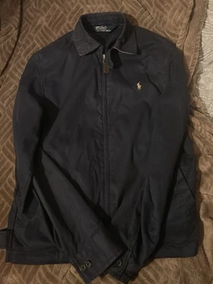 Mörkblå jacka från Polo Ralph Lauren - Klassisk mörkblå jacka från Polo Ralph Lauren med dragkedja framtill och ikonisk broderad logga på bröstet. Jackan har krage, långa ärmar och knappar vid ärmslut och midja. Perfekt för dig som gillar stilren och tidlös design.