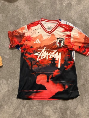 Japan x Stüssy x Adidas tröja - Unik sporttröja i samarbete mellan Japan, Stüssy och Adidas. Tröjan har ett färgstarkt rött och svart landskapsmotiv med japansk pagod, bro och körsbärsblommor. Stüssy-logga framtill, Japan-emblem och Adidas-detaljer. Perfekt för dig som gillar streetwear och sport.