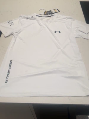 Vit Under Armour t-shirt  - Vit t-shirt från Under Armour med logga på bröstet och tryck längs sidan. Tillverkad i lätt och funktionell polyester, perfekt för träning. Klassisk passform med korta ärmar och rund halsringning.