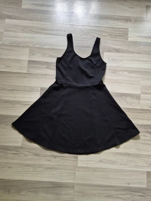 Svart ärmlös kortklänning - En svart, ärmlös kortklänning med rundad halsringning och utställd kjol. Klänningen har en enkel och stilren design, perfekt för att matcha med olika accessoarer. Materialet känns mjukt och följsamt mot kroppen.