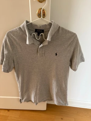 Grå pikétröja från Polo Ralph Lauren - Klassisk grå pikétröja från Polo Ralph Lauren med korta ärmar och krage. Tröjan har en liten broderad logga på bröstet och är tillverkad i mjuk bomull. Perfekt för en clean och stilren look. Pikén är aldrig använd säljer för att jag inte tycker om den.