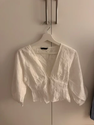 Vit broderad blus med v-ringning - Söt vit blus med broderade detaljer och v-ringning. Blusen har trekvartsärm och smockad midja som ger en feminin siluett. Perfekt att styla med jeans eller kjol nu inför sommaren! Toppen är aldrig använd passar också en mindre M!🌸🩷