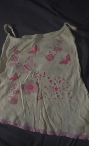 Vit och rosa linnetopp med fjärilar - Söt linnetopp i vitt med rosa detaljer, dekorerad med tryck av fjärilar och blommor framtill. Toppen har smala axelband och en ljusrosa kant längst ner. Perfekt för en somrig look.