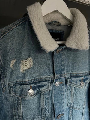 Jeansjacka med teddykrage - Ljusblå jeansjacka från Jack & Jones med vit teddykrage. Jackan har slitna detaljer och klassisk knappstängning framtill. Använd endast ett fåtal gånger, storlek M💫
