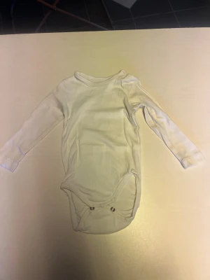 Vit långärmad body i bomull - En enkel och mjuk vit långärmad body med rund hals och tryckknappar i grenen. Perfekt basplagg som är lätt att matcha och skön att bära. Tillverkad i bomull för en bekväm känsla hela dagen.