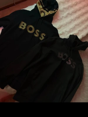 Svart hoodie från BOSS med logga - Svart hoodie från BOSS med stor logga framtill i spegeleffekt. Klassisk modell med huva och dragsko samt känguruficka. Mjuk bomullsblandning och ribbade muddar vid ärmslut och nederkant. Perfekt för en stilren och trendig look. stl L i båda slutsålda! cond 9/10 knappt använda! köpta  på johnells