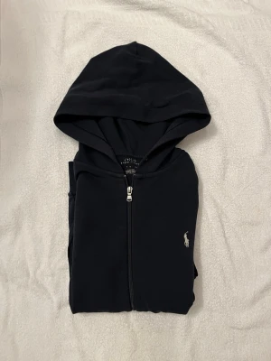 Ralph Lauren zip hoddie  - Klassisk mörk blå hoodie från Ralph Lauren med dragkedja och huva. Ikoniska lilla polospelaren broderad på bröstet. Perfekt för en avslappnad och stilren look. Mjukt material som känns skönt mot huden.