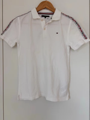 Vit pikétröja Tommy Hilfiger - Vit pikétröja från Tommy Hilfiger med klassisk krage och knappar framtill. Små logodetaljer på bröstet och snygga band med flaggor i rött, vitt och blått längs axlarna. Kortärmad. Strlk 152.