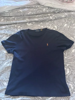 Mörkblå t-shirt från Polo Ralph Lauren - Säljer en mörkblå t-shirt från Polo Ralph Lauren med klassisk passform och korta ärmar. T-shirten har det ikoniska broderade logotypen på bröstet och är gjord i mjuk bomull. Perfekt för dig som gillar stilrena basics med en touch av premium.