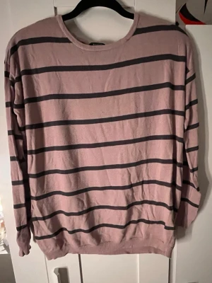 Rosa tröja med svarta ränder - En stilren rosa tröja med svarta horisontella ränder. Tröjan har rund halsringning, långa ärmar och ribbade muddar vid ärmslut och nederkant. Perfekt för en avslappnad och trendig look.