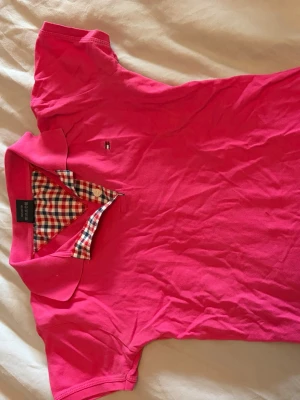 Rosa pikétröja Tommy Hilfiger S - Snygg rosa pikétröja från Tommy Hilfiger med klassisk krage och rutigt tyg på insidan av knäppningen. Liten logga på bröstet och korta ärmar. Perfekt för en avslappnad men stilren look. Vet inte om den är äkta 