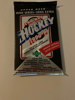 Upper Deck NHL Hockeykort 1990-91 - Ett oöppnat paket med 12 samlarkort från Upper Deck NHL Hockey 1990-91, French Edition. Förpackningen är svart med stora blå, röda och vita tryck. Perfekt för dig som gillar retro och samlarkort från hockeyvärlden.