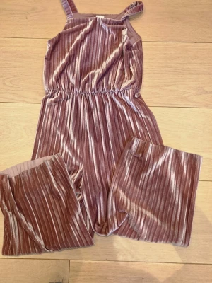 Rosa plisserad jumpsuit från Lindex - Supersnygg rosa jumpsuit från Lindex i plisserat sammetstyg. Den har smala axelband, resår i midjan och vida ben. Perfekt för dig som vill ha en trendig och bekväm look med lite extra glans.