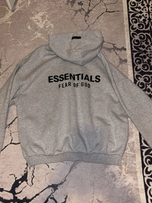 Grå Essentials Fear of God hoodie - Snygg grå hoodie från Fear of God Essentials med svart tryck både fram och bak. Oversized passform, ribbade muddar och stor huva. Perfekt för dig som gillar streetwear och vill ha en stilren look. Materialet är mjukt och skönt, troligtvis bomullsblandning. Pris går att diskutera. Kolla profil för även svart färg samt byxor till den.