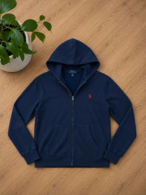 Mörkblå hoodie från Polo Ralph Lauren - Mörkblå hoodie med dragkedja från Polo Ralph Lauren. Klassisk modell med huva, kängurufickor och röd broderad logga på bröstet. Tillverkad i mjuk bomullsblandning för en skön känsla och stilren look. Tyget har gått upp lite på ett ställe men det syns knappt 