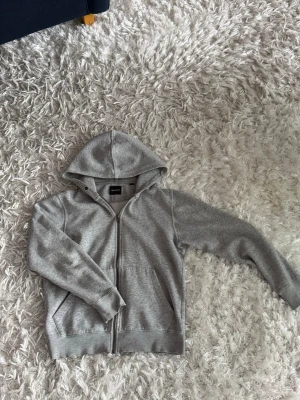 Grå hoodie med dragkedja Jack & Jones - Snygg grå hoodie från Jack & Jones med dragkedja framtill och två fickor. Mjuk insida och klassisk huva. Perfekt för en avslappnad stil och enkel att matcha med jeans eller joggers.