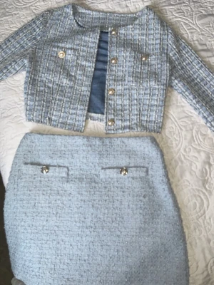 Ljusblått tweed set med kjol och jacka - Snyggt tvådelat set i ljusblå tweed med kort jacka och matchande kjol. Jackan har guldiga knappar och rund halsringning, medan kjolen har dekorativa fickor framtill med knappar. Perfekt för en chic och trendig look.