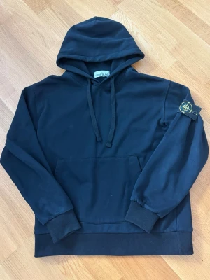 Svart hoodie från Stone Island - Svart hoodie från Stone Island med klassisk huva och dragsko. Har en stor magficka och den ikoniska loggan på vänster ärm. Tillverkad i mjuk bomull, perfekt för en avslappnad och stilren look. Din sitter som S/M.