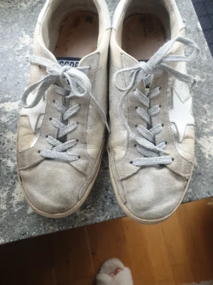 Beige och vita Golden Goose sneakers - Snygga sneakers från Golden Goose i beige mocka och vitt läder med den ikoniska stjärnan på sidan. Skorna har silvriga skosnören och en klassisk låg modell. Perfekta för dig som gillar en avslappnad och trendig stil.