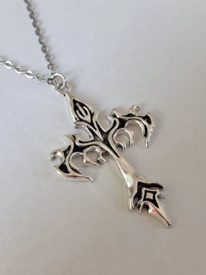 Silverfärgat halsband med korshänge - Snyggt halsband med ett stort korsformat hänge i silverfärgad metall. Hänget har en unik, ornamenterad design med svarta detaljer som ger en cool och edgy känsla. Perfekt accessoar för dig som gillar statement-smycken och vill sticka ut. Materialet rostfritt stål. 