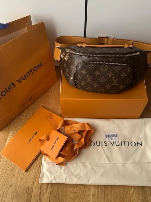 Louis Vuitton monogram magväska - Snygg magväska från Louis Vuitton i klassiskt brunt monogrammönster med guldiga detaljer och justerbart beige läderband. Väskan har flera fack med dragkedja och är tillverkad i canvas och läder. Perfekt accessoar för en trendig look.