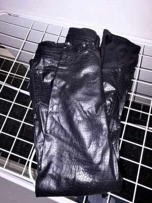 Svarta jeans med ormskinnsmönster - Snygga svarta jeans med glansig finish och ormskinnsmönster. Jeansen har klassisk femficksdesign och raka ben. Perfekta för dig som vill sticka ut med en trendig look. Materialet ser ut som konstläder med struktur.