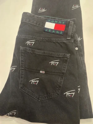 Svarta jeans från Tommy Jeans - Snygga svarta jeans från Tommy Jeans med broderade logotyper över hela byxan. Klassisk femficksmodell med raka ben och normal midja. Jeansen har en patch med Tommy Jeans-flaggan bak i midjan. Tillverkade i bomull. Storlek W 33 och L 32🥰