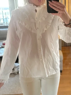 Vit volangblus med spetsdetaljer - Söt vit blus med volang och spets längs bröstet och kragen. Blusen har knäppning framtill, hög krage med spets och långa ärmar med spets vid ärmslut. Perfekt för en romantisk och trendig look. Storlek 44 men sitter som M