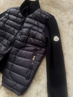 Marinblå cardiganjacka från Moncler - Snygg marinblå cardiganjacka från Moncler med quiltad framsida i glansigt material och stickade ärmar. Jackan har dragkedja framtill, två fickor med dragkedja och Moncler-logga på ärmen. Perfekt för lager-på-lager och streetstyle.