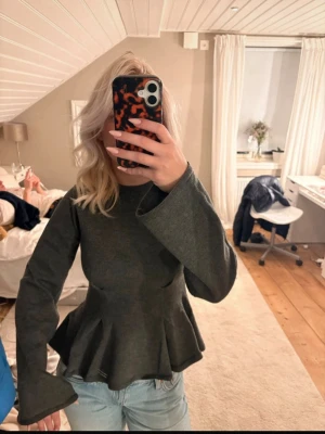 Grå peplumtop med vida ärmar - Snygg grå topp med peplumskärning och vida långa ärmar. Toppen har en rund halsringning och är tillverkad i ett mjukt material som ger en bekväm passform. Perfekt att styla med jeans för en trendig look.