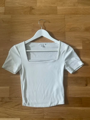 Vit croppad topp med fyrkantig ringning - Säljer en vit croppad topp från New yorker med fyrkantig ringning och korta ärmar. Toppen har en slim passform. Perfekt till jeans eller kjol för en clean look.