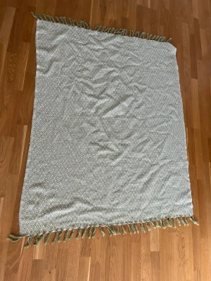 Grön filt med diamantmönster och fransar - Säljer en grön filt med vitt diamantmönster och fransar längs kanterna. Filten är rektangulär och gjord av mjukt tyg, perfekt som inredningsdetalj eller att ha i soffan. Enkel och stilren design som passar i de flesta hem.