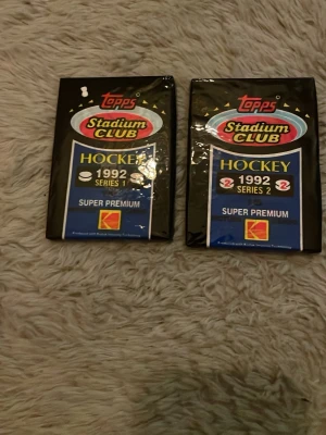 Topps Stadium Club Hockeykort 1992 - Två oöppnade paket Topps Stadium Club hockeykort från 1992, serie 1 och serie 2. Svart och blå förpackning med färgglad logga. Perfekt för samlare av sportkort och nostalgi från 90-talet.