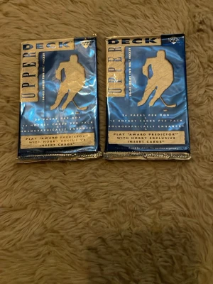 Upper Deck hockeykort boosterpackar - Två oöppnade boosterpackar med hockeykort från Upper Deck. Förpackningarna är blå och guld med holografiska detaljer och innehåller samlarkort med hockeytema. Perfekt för dig som gillar att samla sportkort eller vill hitta exklusiva inserts.